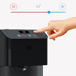 A hand presses the cold water button on a black Brio water dispenser. The screen above shows a temperature of 62 degrees, with hot and cold icons and a blue slider indicating a cold setting.